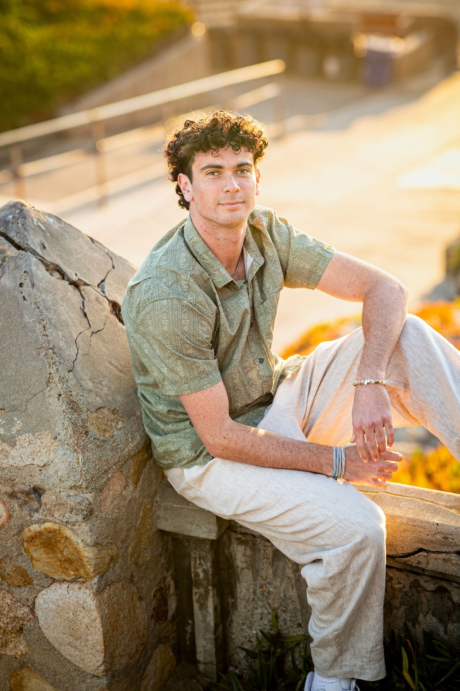 A person in casual attire sits casually on a stone wall during golden hour lighting.
