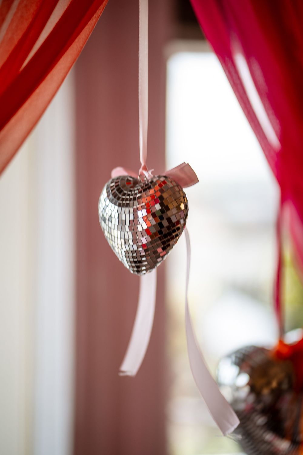 Shimmering silver disco ball hearts hang from red ribbons against a soft blurred background.