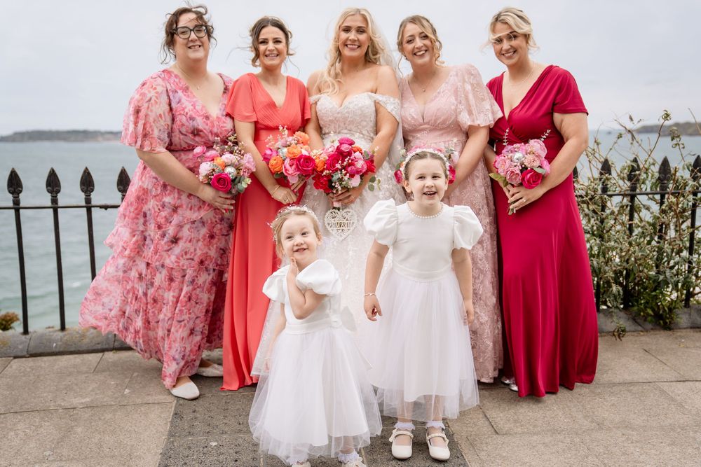 Wedding party in pink and coral dresses pose with two flower girls in white on a seaside terrace.
