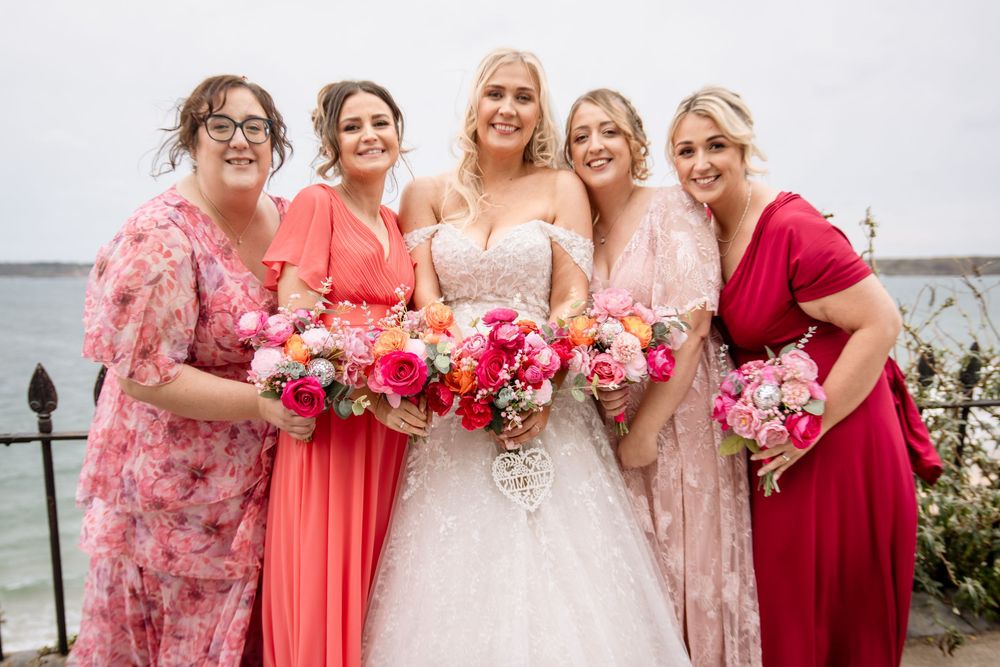 A wedding party in coral and red dresses poses together holding vibrant pink and white floral bouquets.