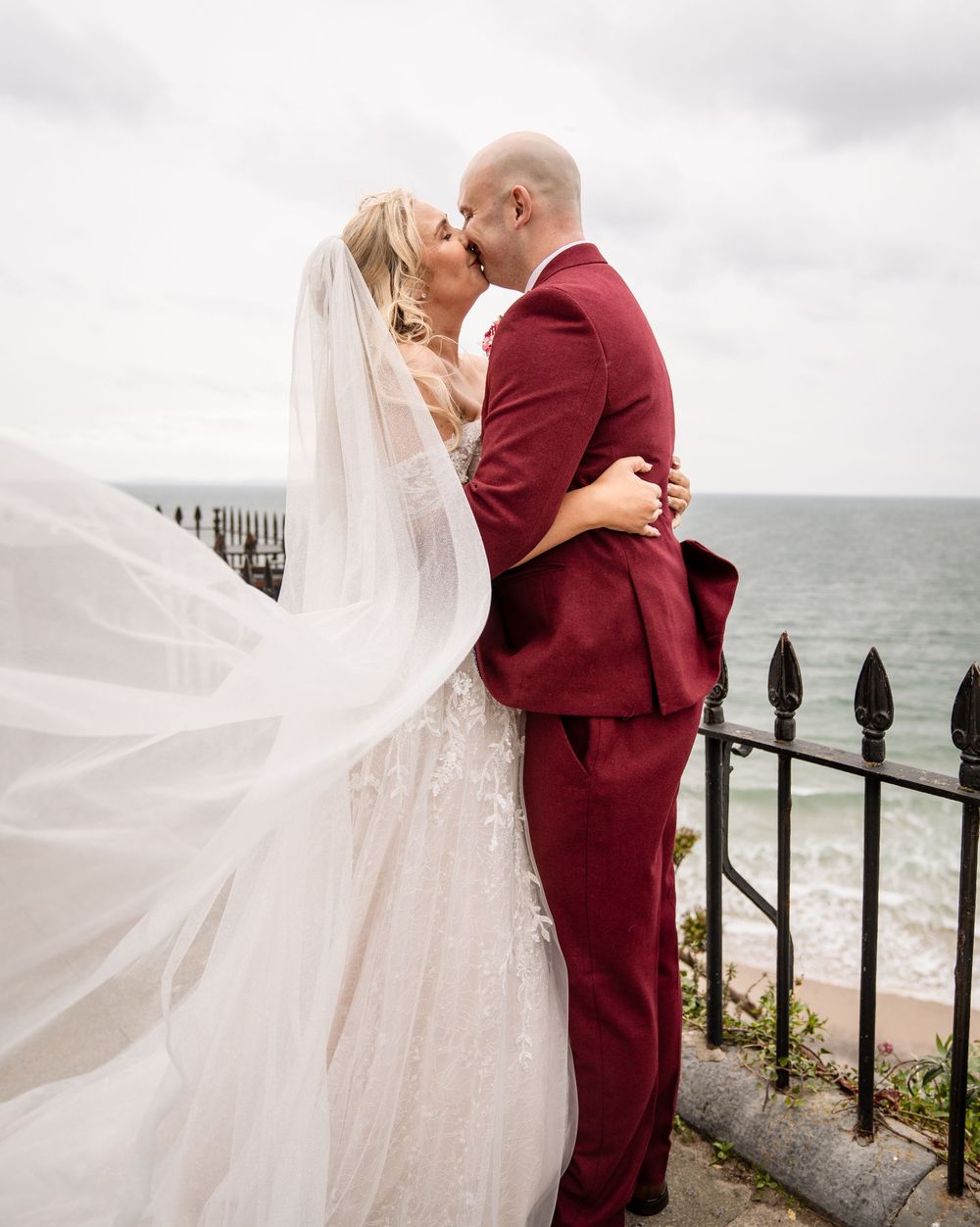 Romantic wedding photos by the ocean with bride in white dress and groom in burgundy suit.