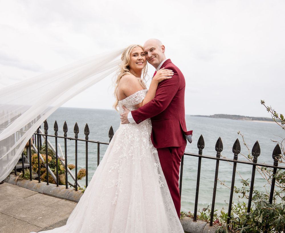 A couple in burgundy and white wedding attire embrace by the ocean on their wedding day.