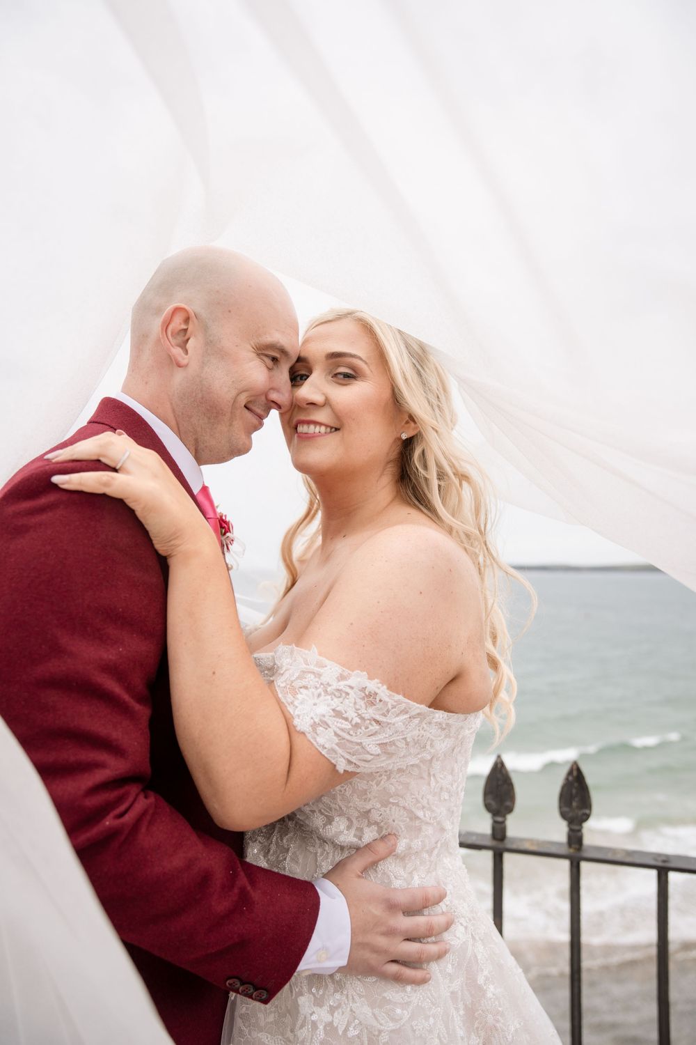 A romantic seaside wedding portrait with a burgundy suit and lace wedding dress against an ocean backdrop.