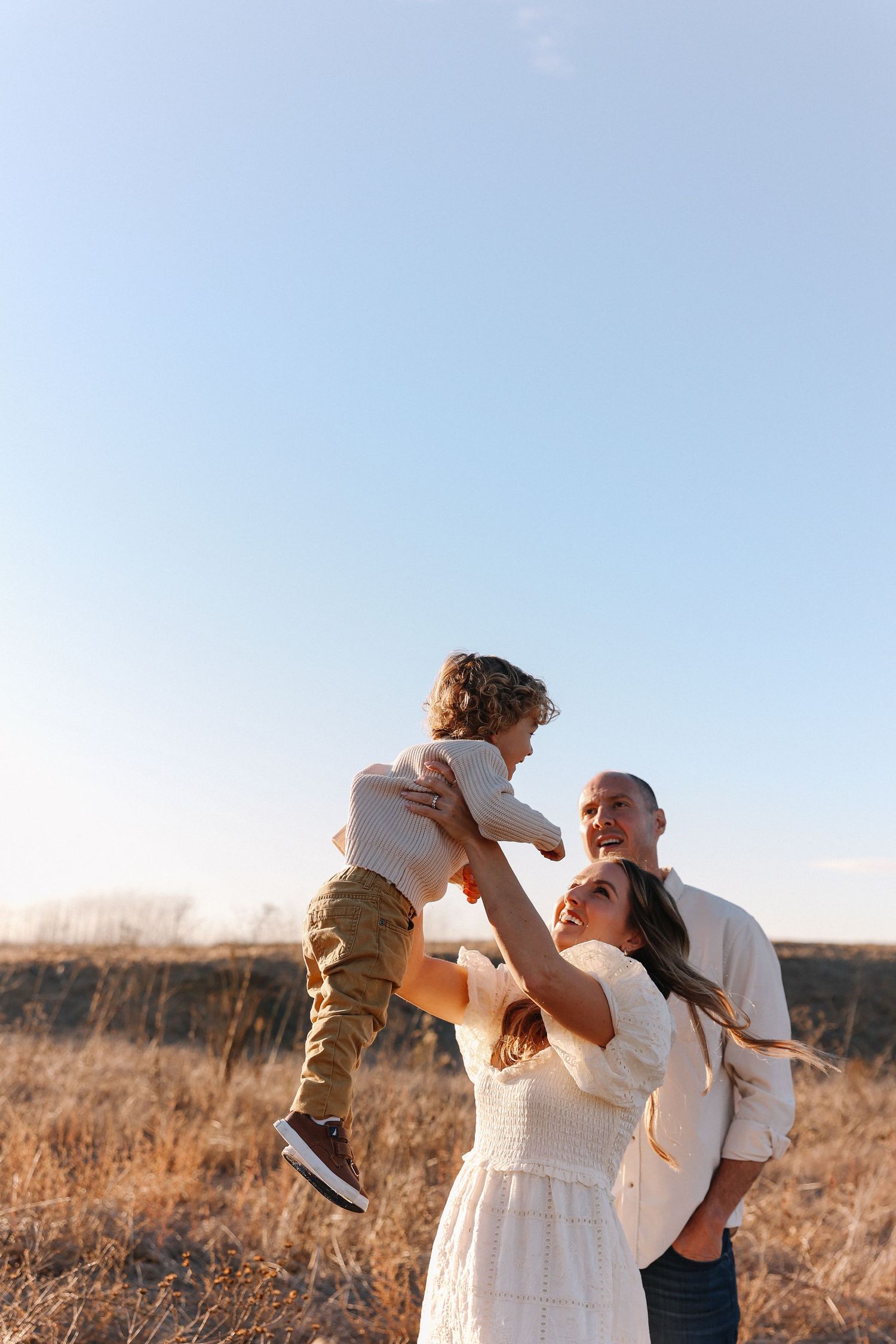 A family shares a joyful moment together in a golden field during sunset, with mom lifting a her toddler up high.