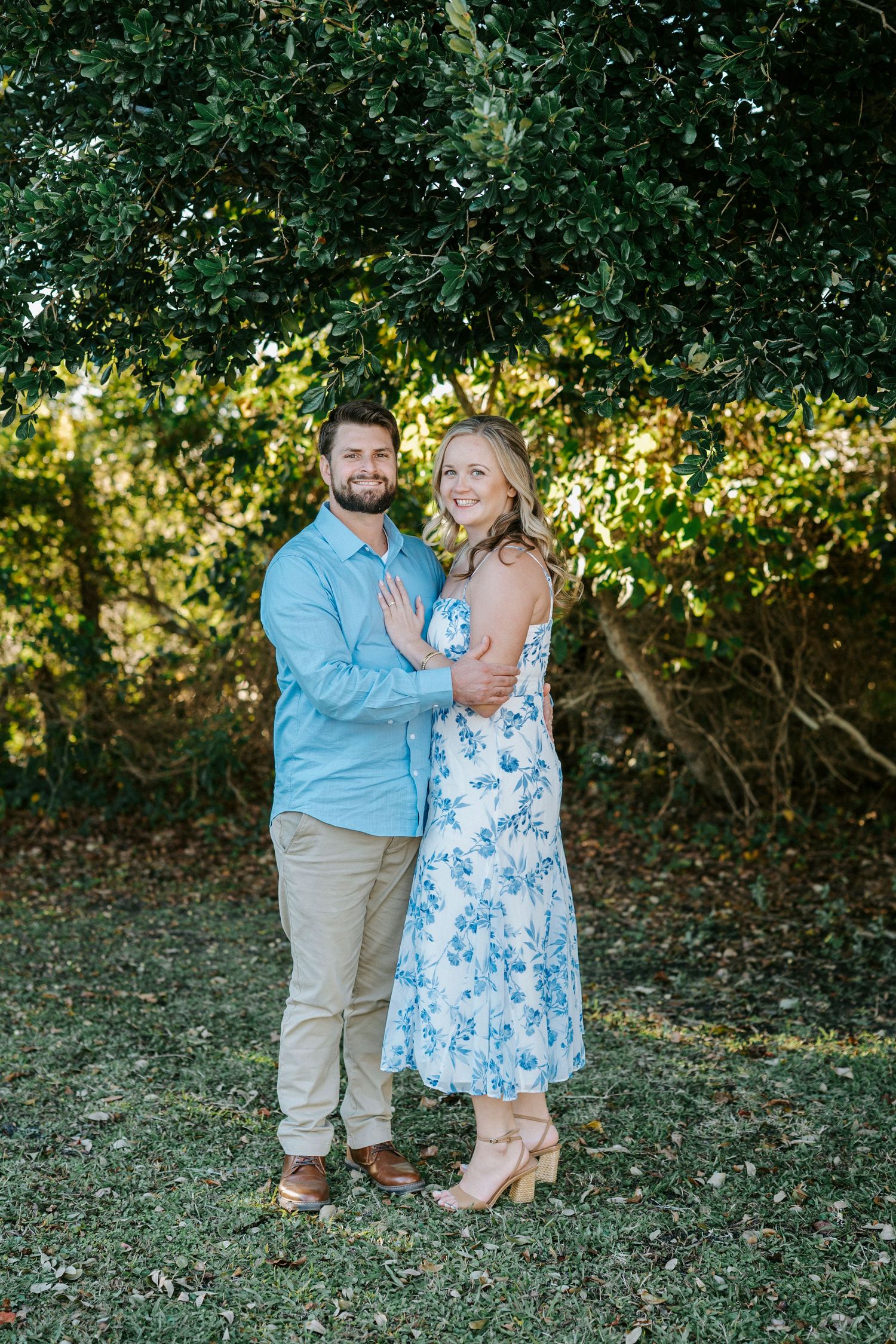 A couple shares tender moments in an outdoor setting wearing coordinated blue and khaki outfits against a leafy backdrop.