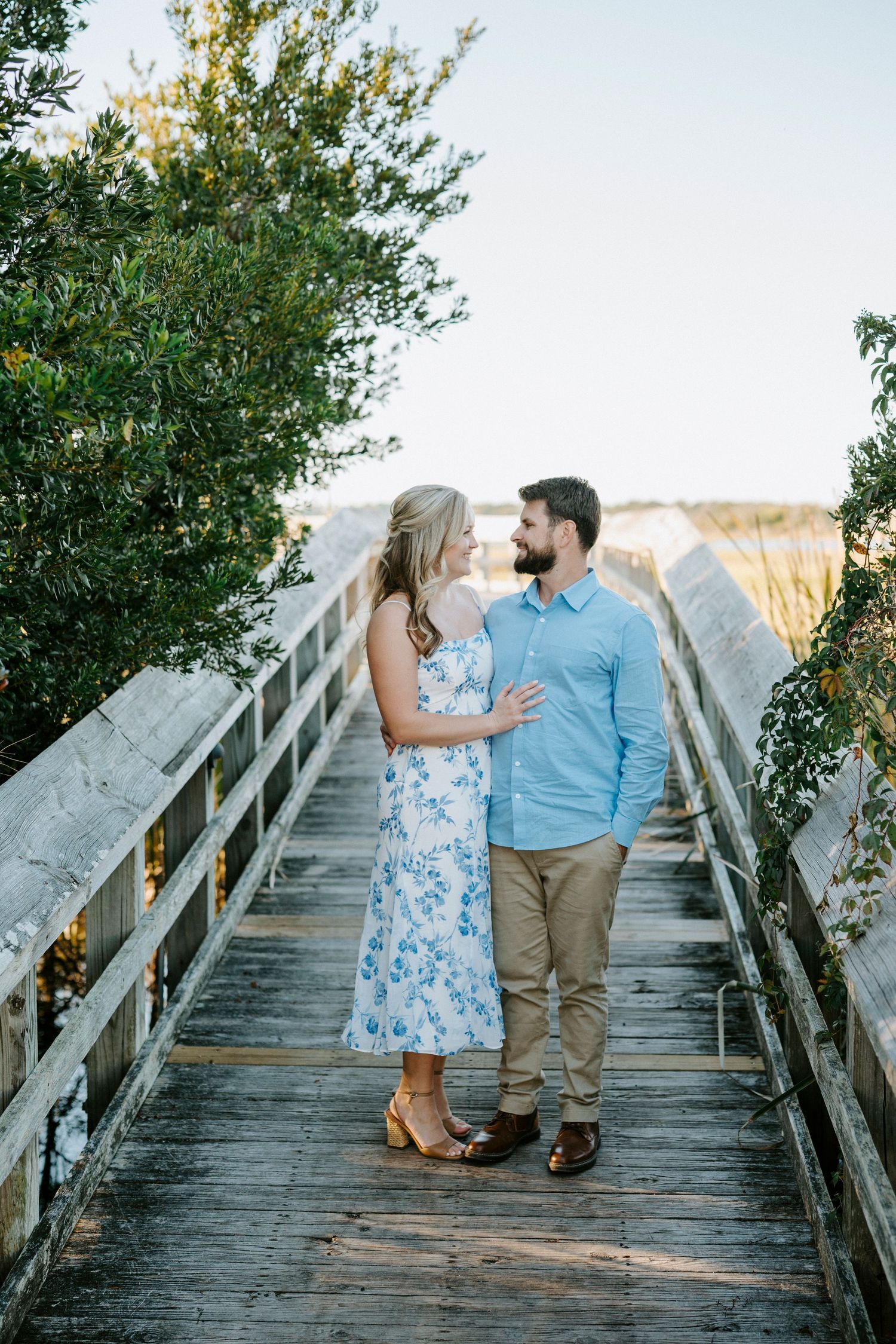 A couple stands lovingly together on a wooden bridge surrounded by greenery during a summer engagement photoshoot.