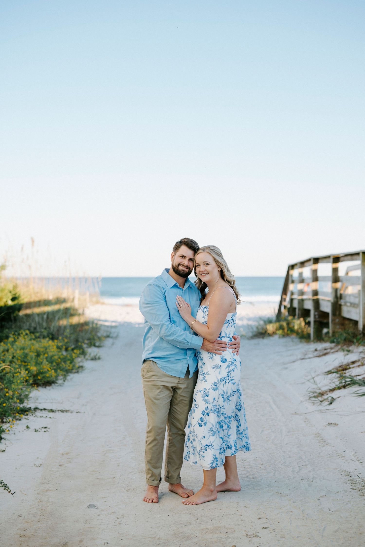 A couple shares a romantic moment walking along a sandy beach path on a sunny afternoon.