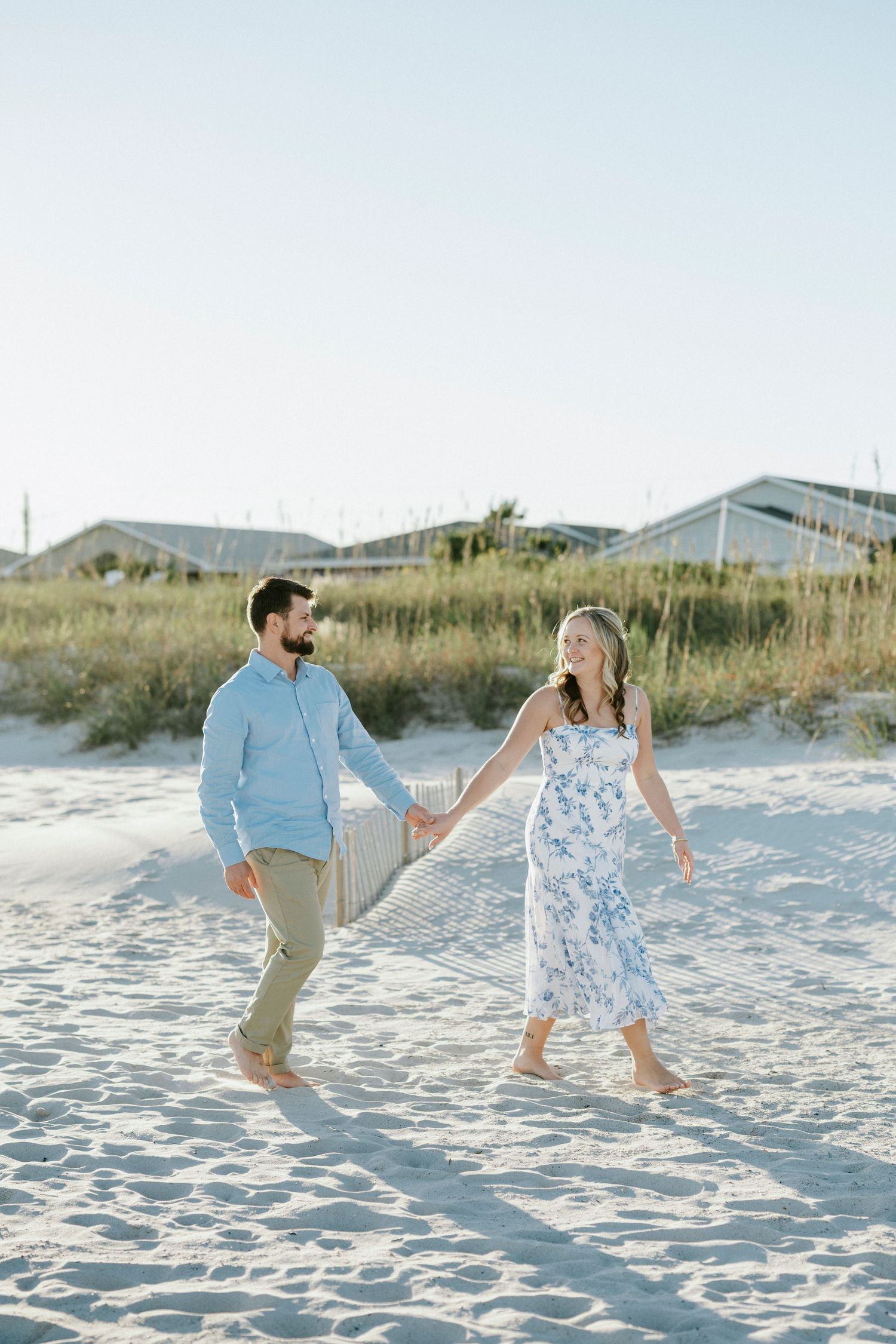 A couple walks hand in hand on a sunny beach with sand dunes and beach houses in the background.