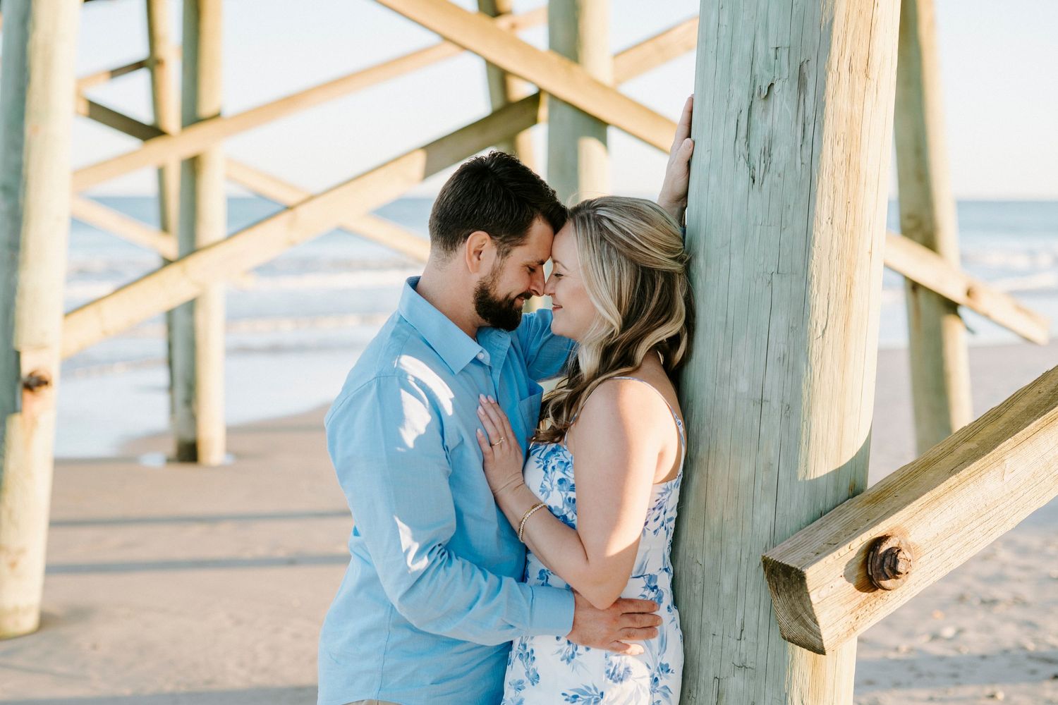 A romantic couple in blue and white outfits share tender moments under a wooden pier at sunset.