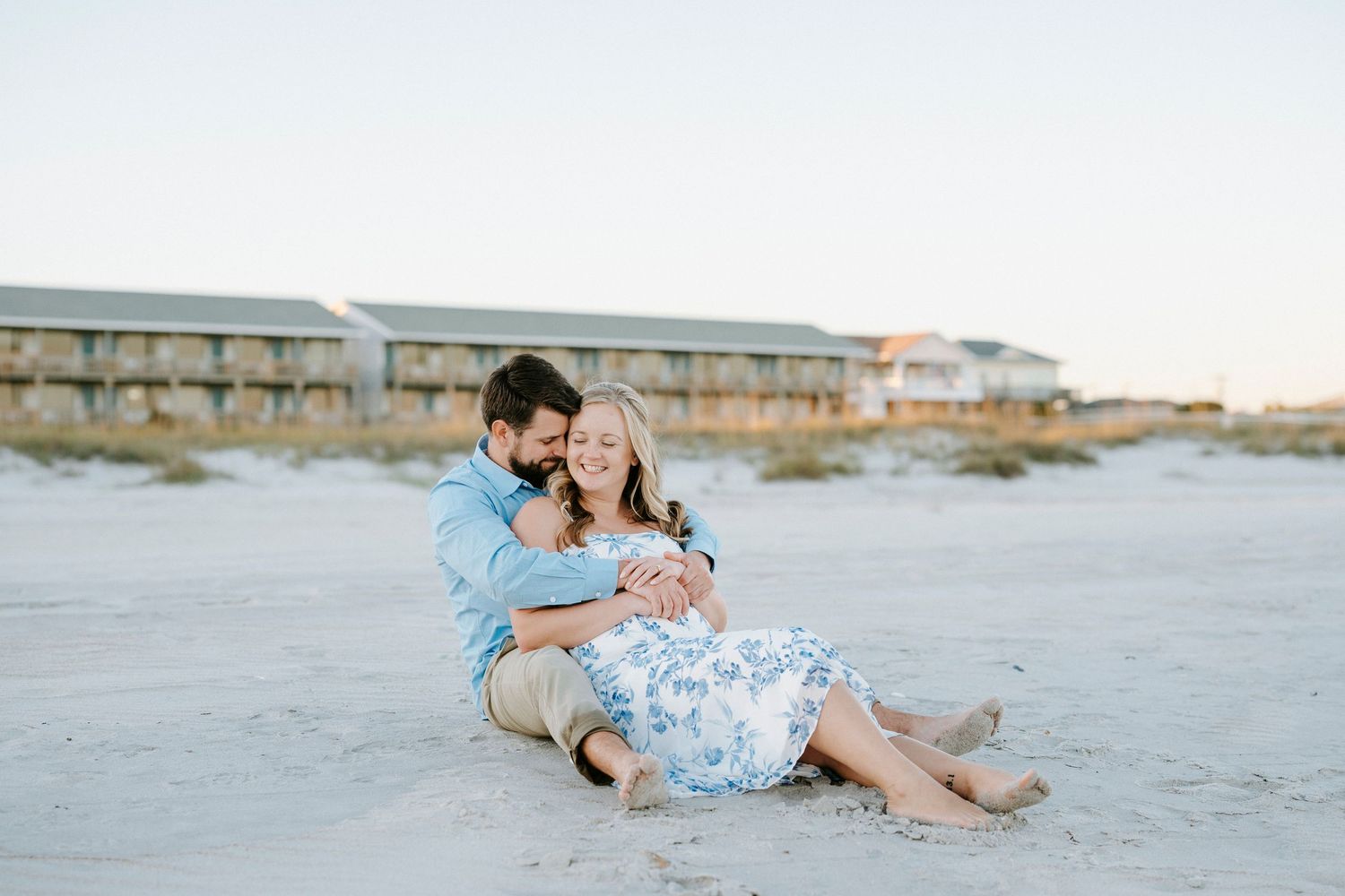 A couple in blue and white outfits share romantic moments on a sandy beach at sunset.
