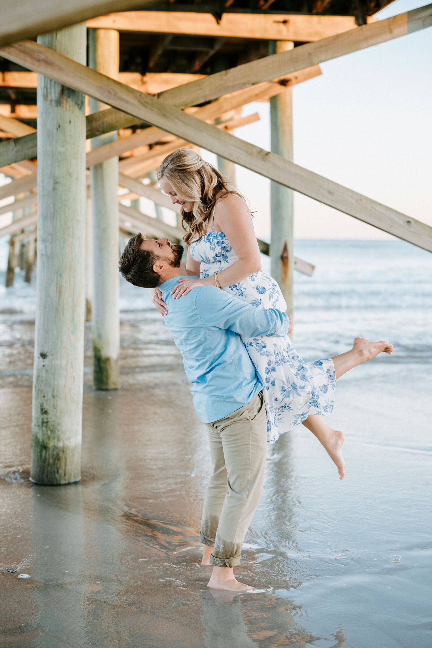 Romantic couple embraces under a beach pier at sunset with waves lapping at their feet.