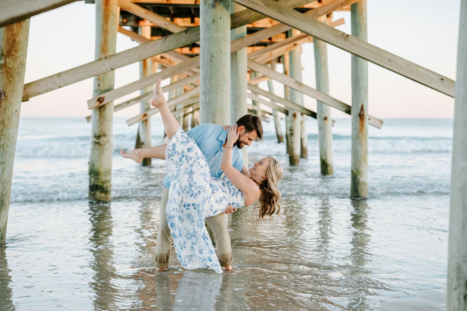 A couple stands in the ocean waves under a wooden pier during sunset creating a romantic silhouette.