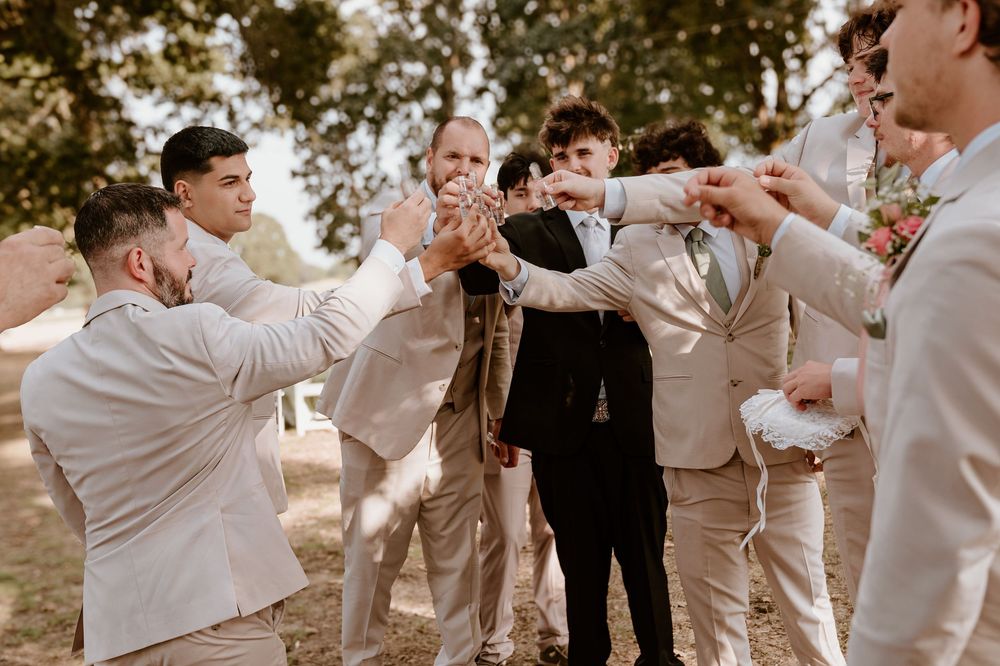 Groom celebrating with groomsmen in tan suits during outdoor wedding party gathering under trees.