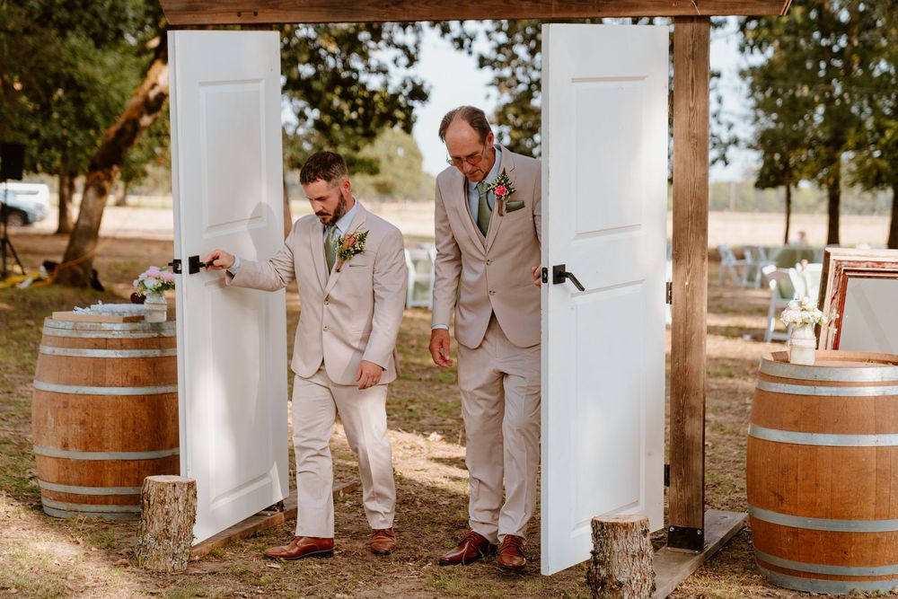 Groom and groomsman in tan suits opening white doors at rustic outdoor wedding venue with barrels.