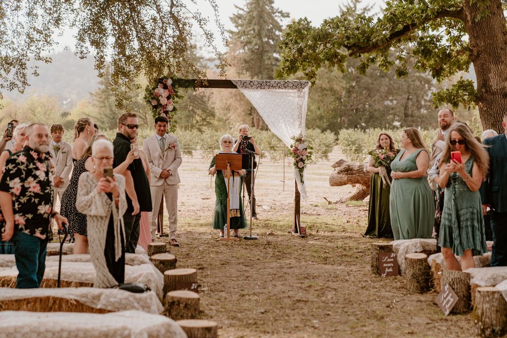 Guests gathering along tree-lined aisle at rustic outdoor wedding ceremony with log seating.