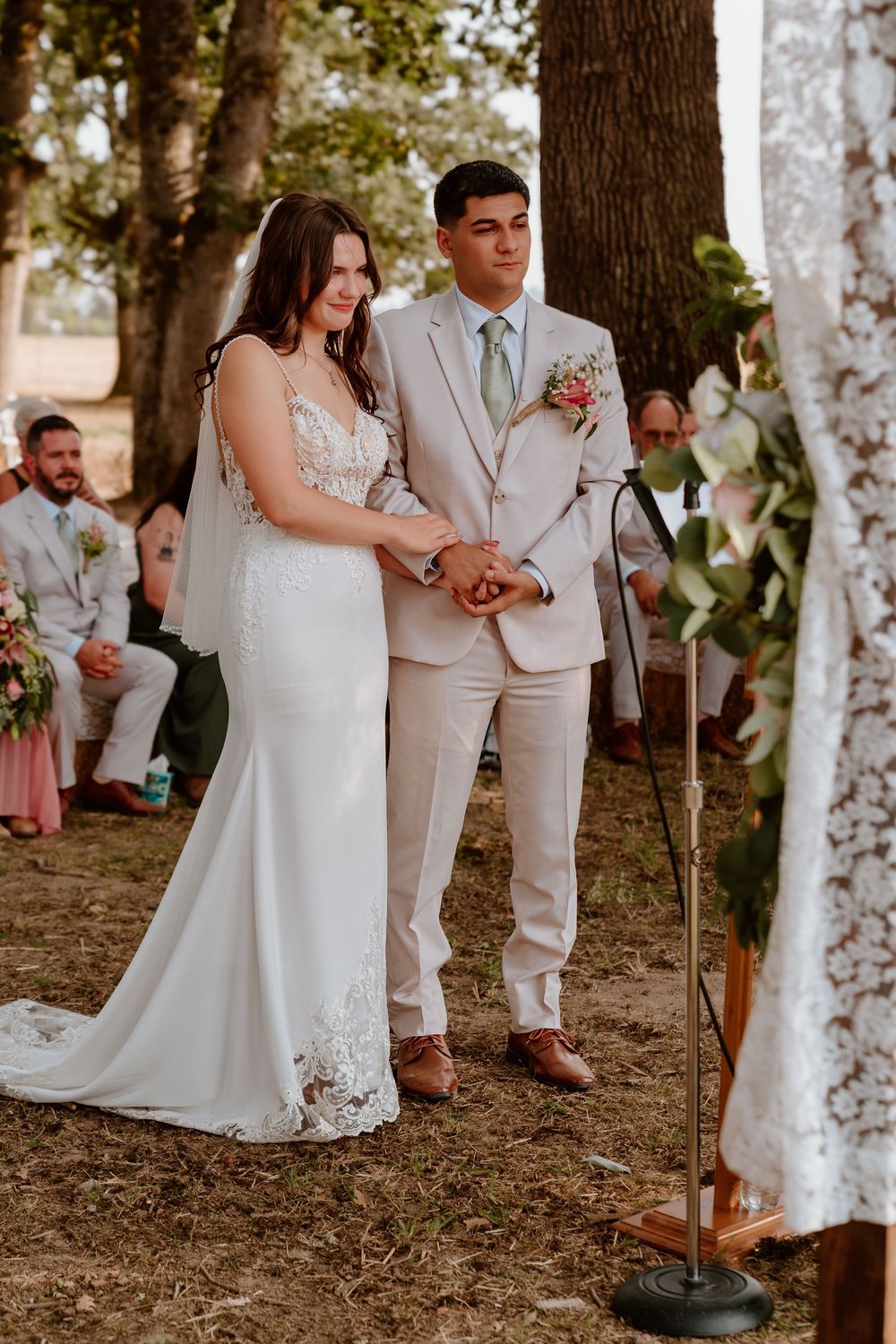 Bride and groom holding hands at outdoor wedding ceremony beneath trees with lace altar decoration.