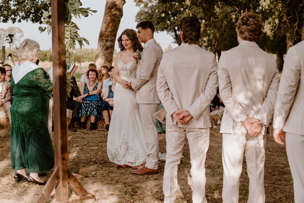 Bride and groom hold hands during outdoor wedding ceremony under trees with guests seated behind.