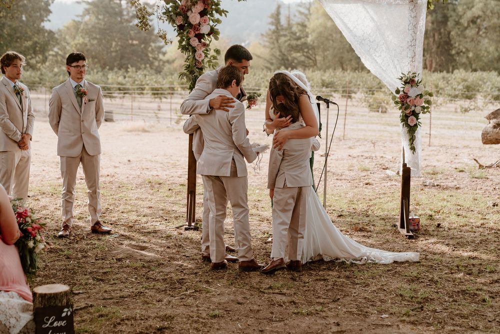 Bride and groom embrace wedding guest in emotional moment at outdoor rustic ceremony altar.
