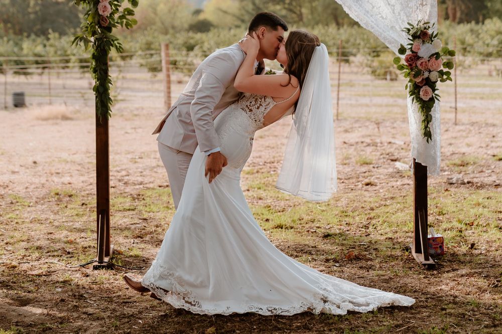Newlyweds share first kiss in front of floral wedding arch at rustic outdoor ceremony.