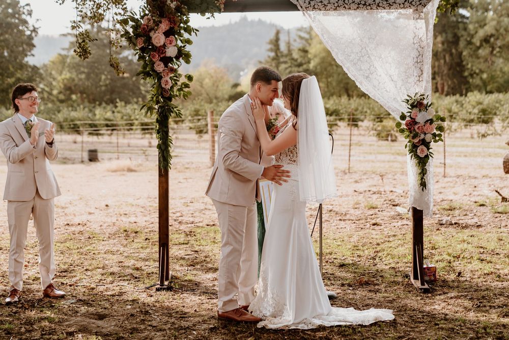 Bride and groom share romantic first kiss under floral arch at sunny outdoor wedding ceremony.