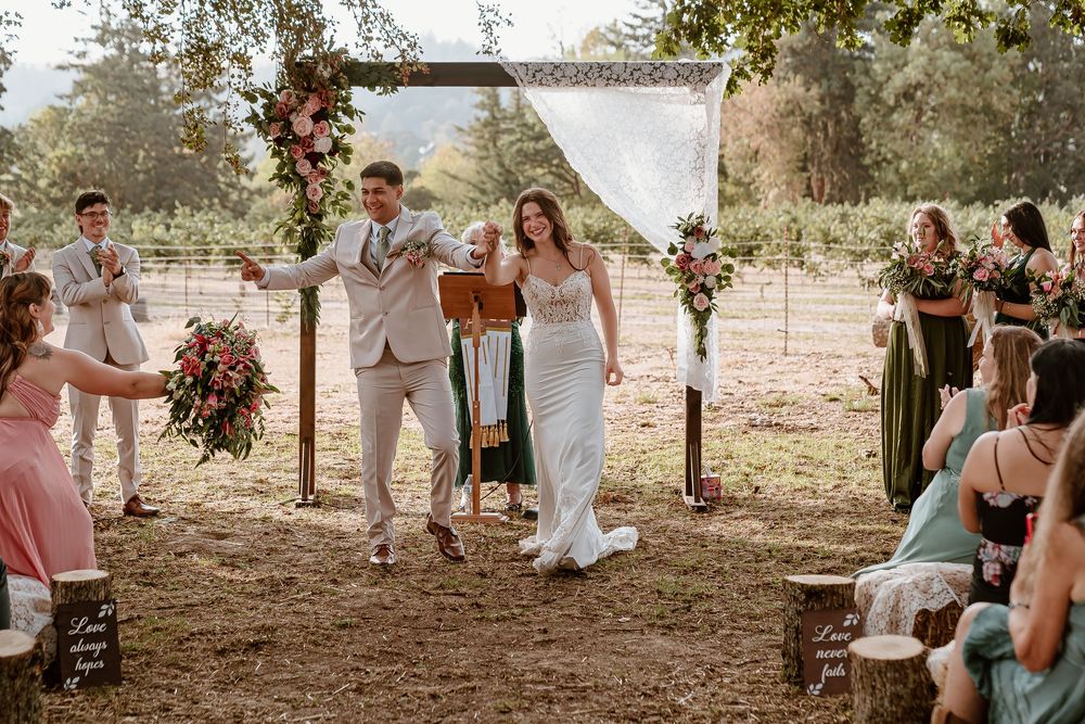 Newlyweds joyfully walk down aisle after ceremony as guests celebrate at outdoor vineyard wedding.