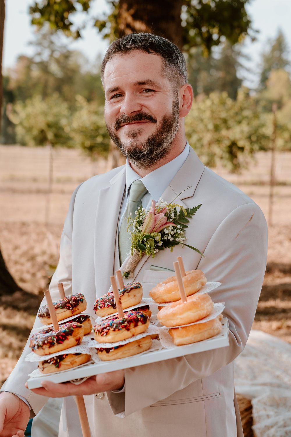 Bearded groomsman in tan suit holds tray of assorted donuts at outdoor wedding reception.
