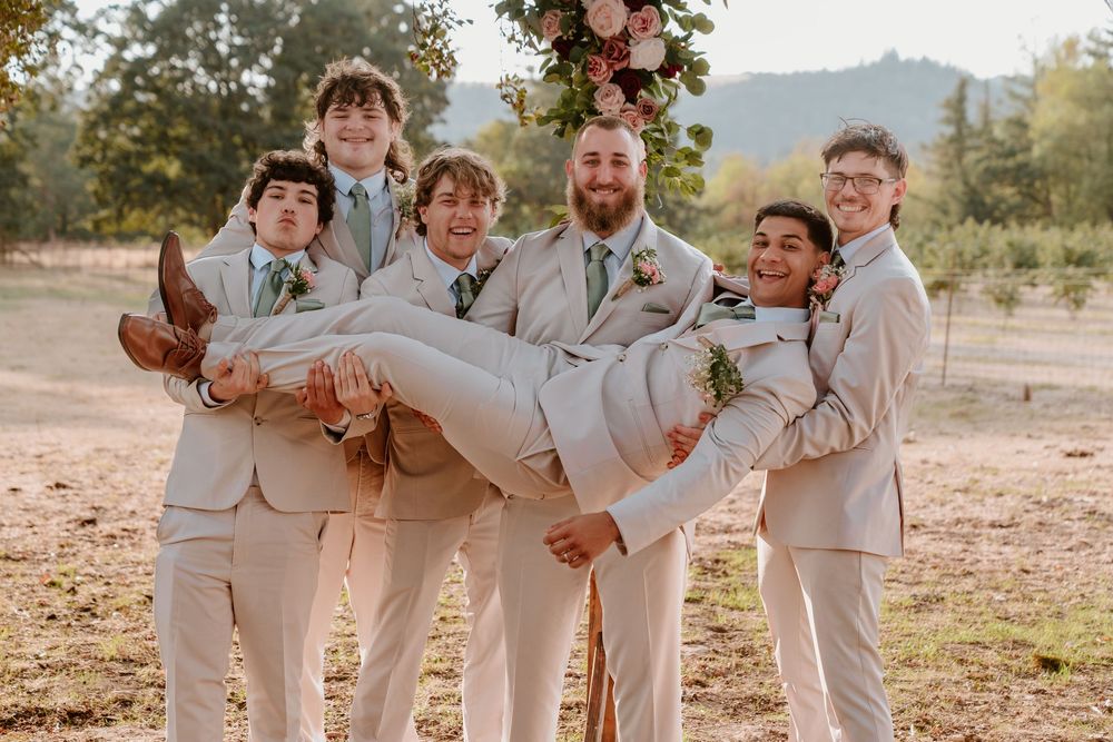 Groomsmen in tan suits playfully lift groom at outdoor wedding celebration near floral arch.