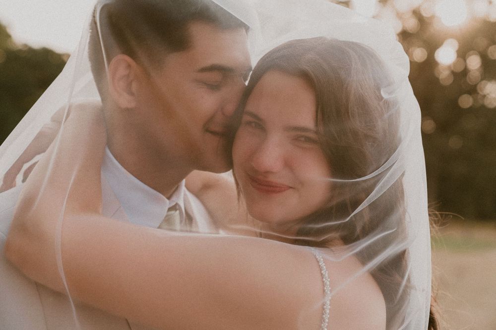Bride and groom share a tender moment under a flowing white veil during their outdoor wedding ceremony.