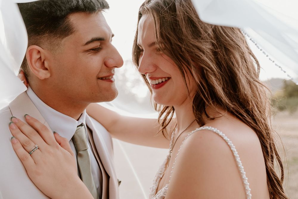 Smiling bride and groom share a joyful moment, with bride wearing engagement ring and groom in white suit.