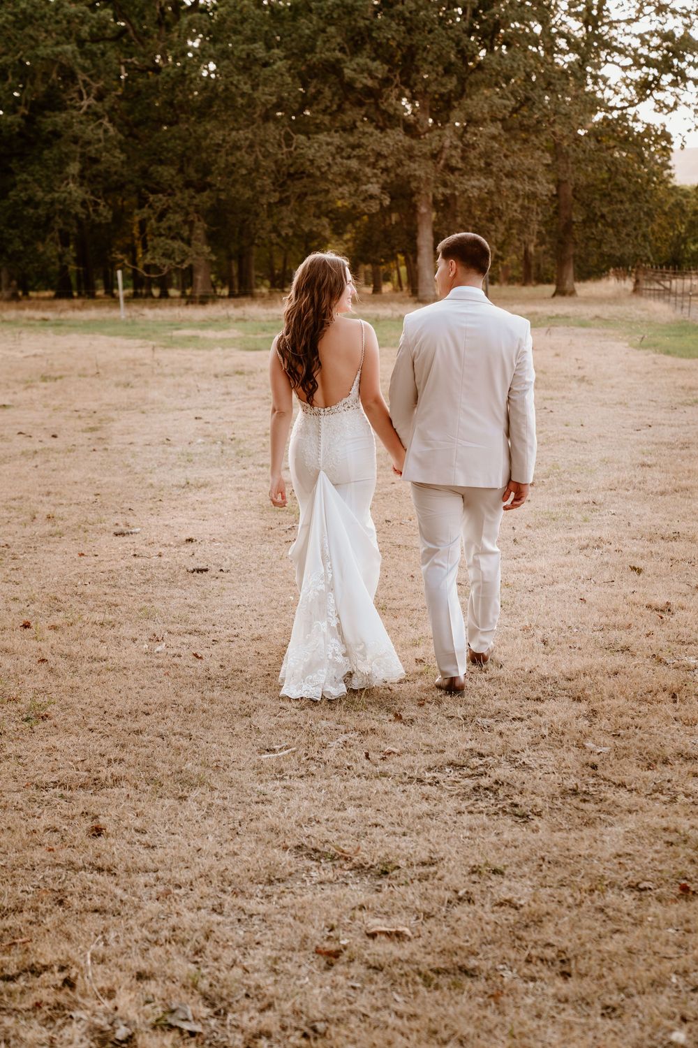 Bride in backless lace gown and groom in tan suit walk hand in hand through a golden field.