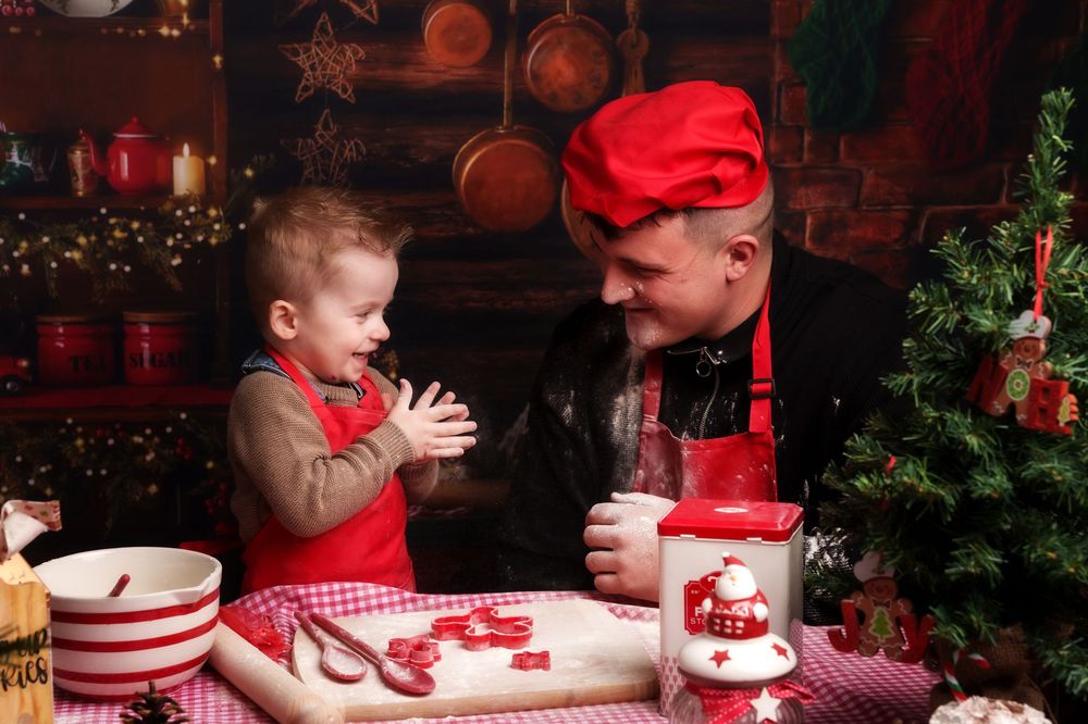 Two people in red holiday outfits decorate Christmas cookies with frosting and sprinkles at a festive table.
