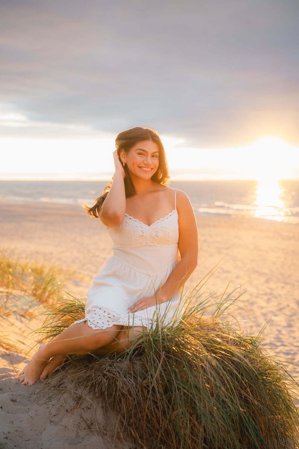 Woman in white dress poses on beach grass during golden hour sunset.