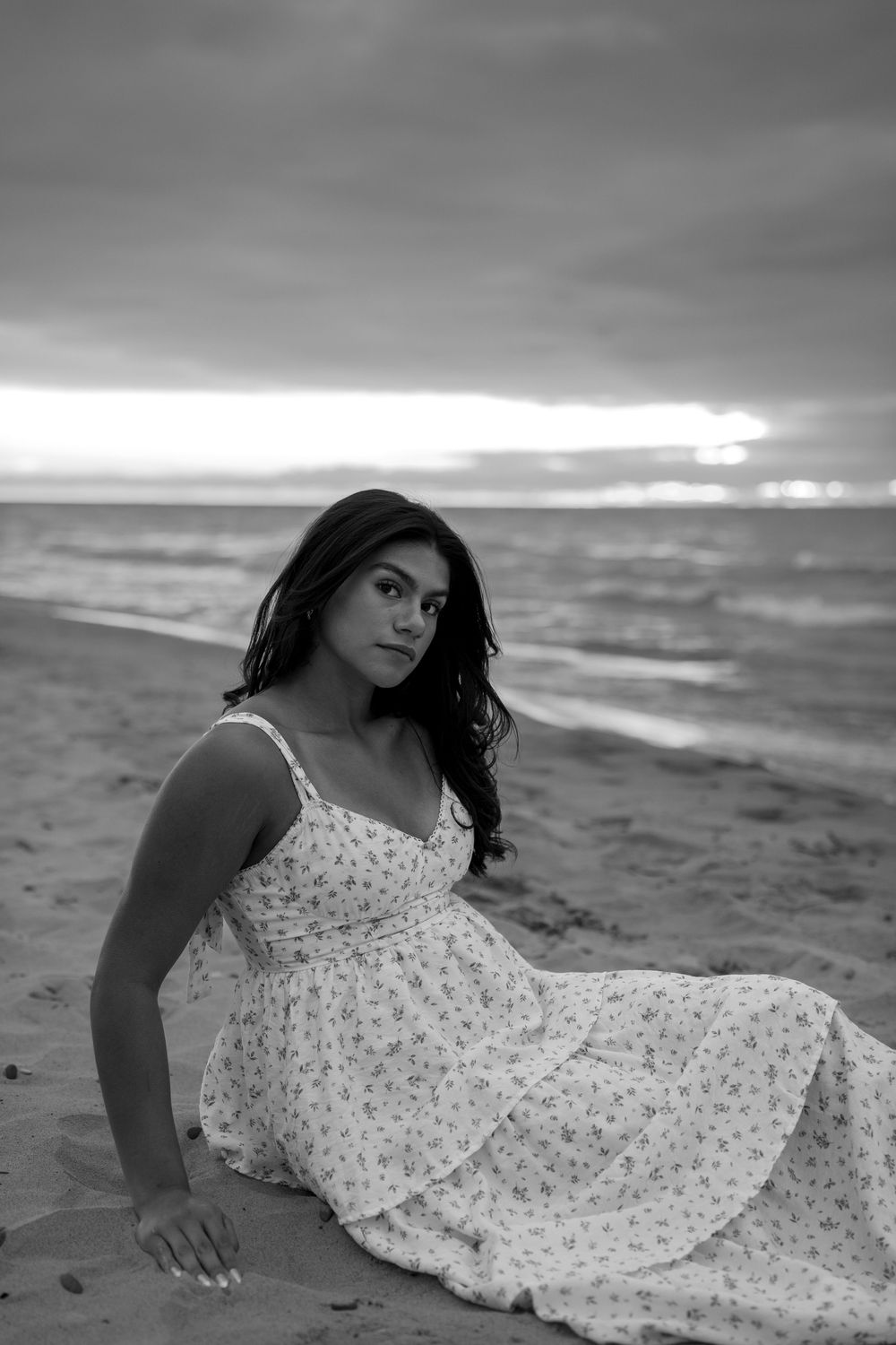 A person in a floral sundress poses gracefully on a beach during sunset in black and white.