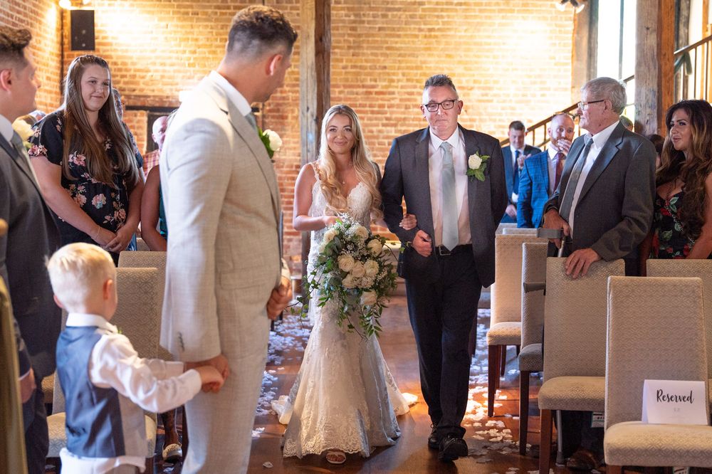 The bride arrives in the aisle as her groom looks back at her, she is in an elegant lace gown and he is wearing a light suit.