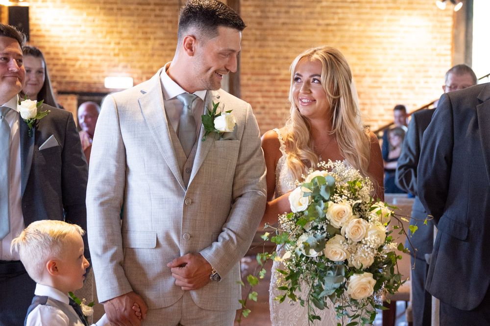 A loving smile to each other as the bride arrives at the top of the aisle, guests in the background are all looking on and smiling.
