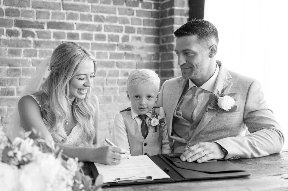 A black and white image of the bride and groom and their young son as they sign their wedding register.
