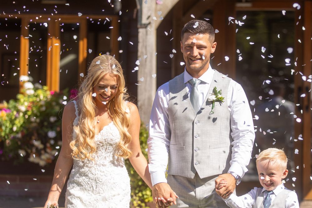 Joyful wedding couple and child celebrate under falling confetti in front of a sunlit garden backdrop of their summer wedding.