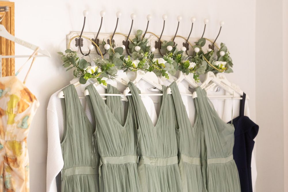 Row of sage green bridesmaid dresses hanging on white hangers against a light coloured wall in the Bridal prep room at Cooling Castle Barn.