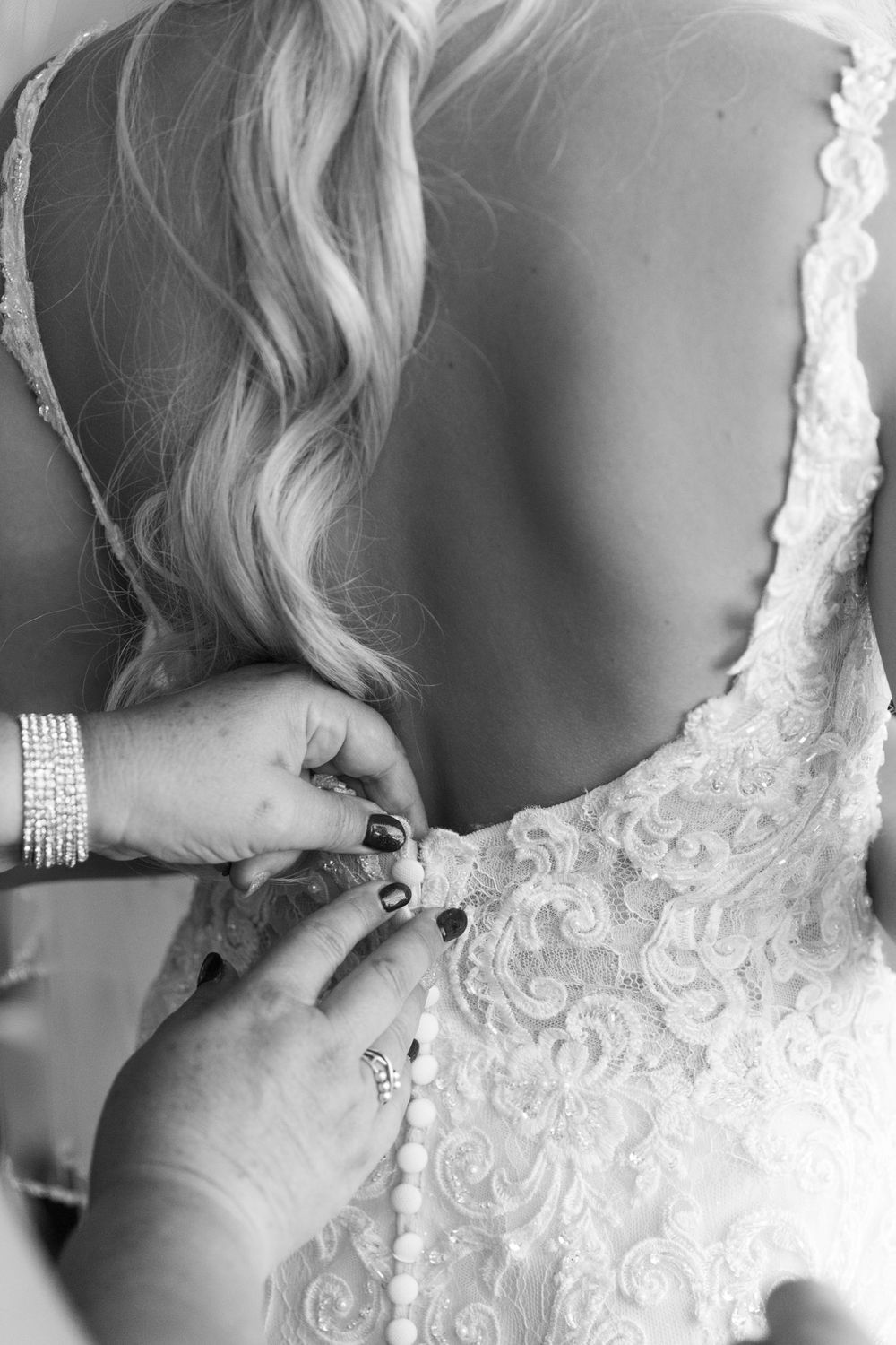 Close-up detail of delicate white lace wedding dress with intricate floral embroidery patterns. The brides mother is fastening the buttons.
