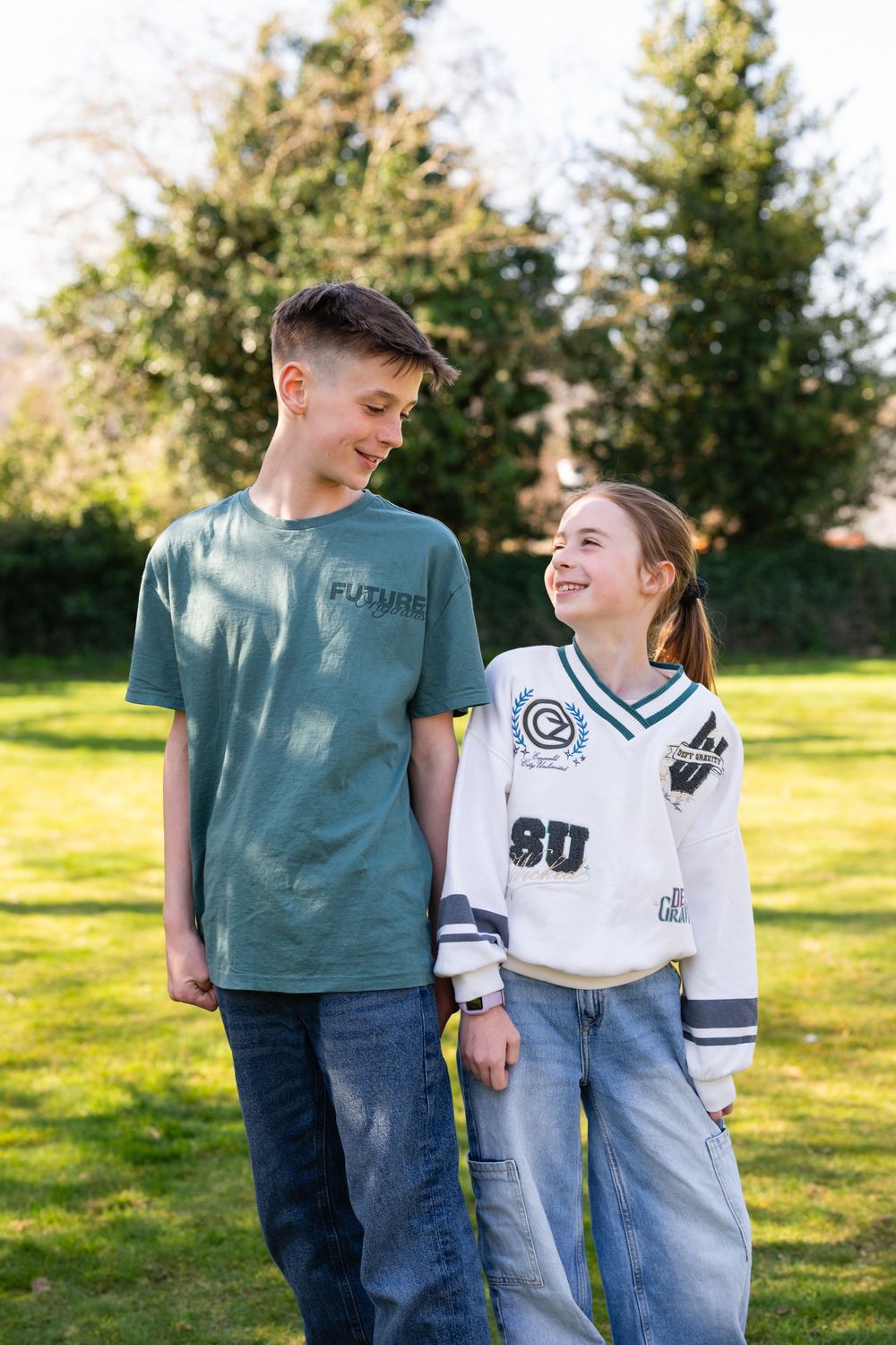 Two young people in casual clothing share a candid moment while standing on grass with evergreen trees in background.