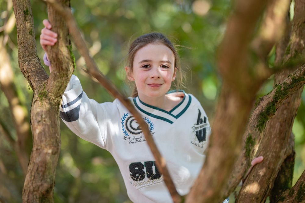 A young person in a white and navy varsity-style sweater gives thumbs up while posing near tree branches.