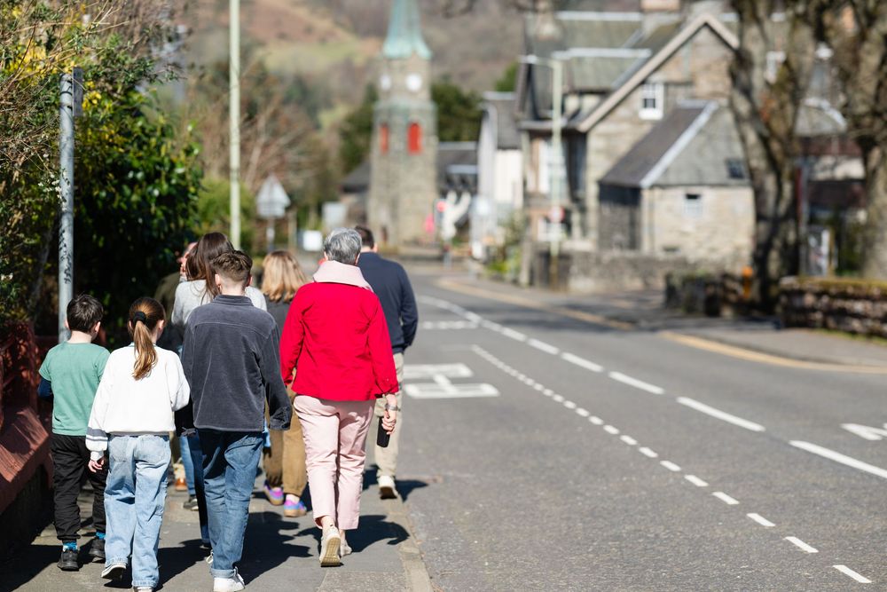 Group of people walking along a village road with church spire visible in the background on a sunny day.