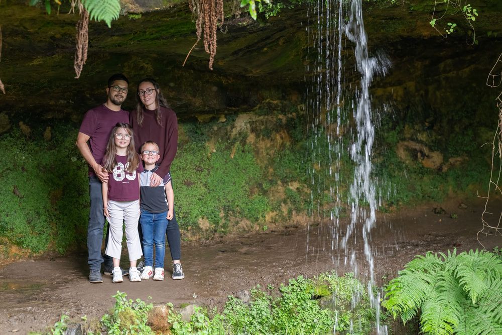 A family poses in front of a mossy rock wall with hanging vines and ferns in a lush outdoor setting.