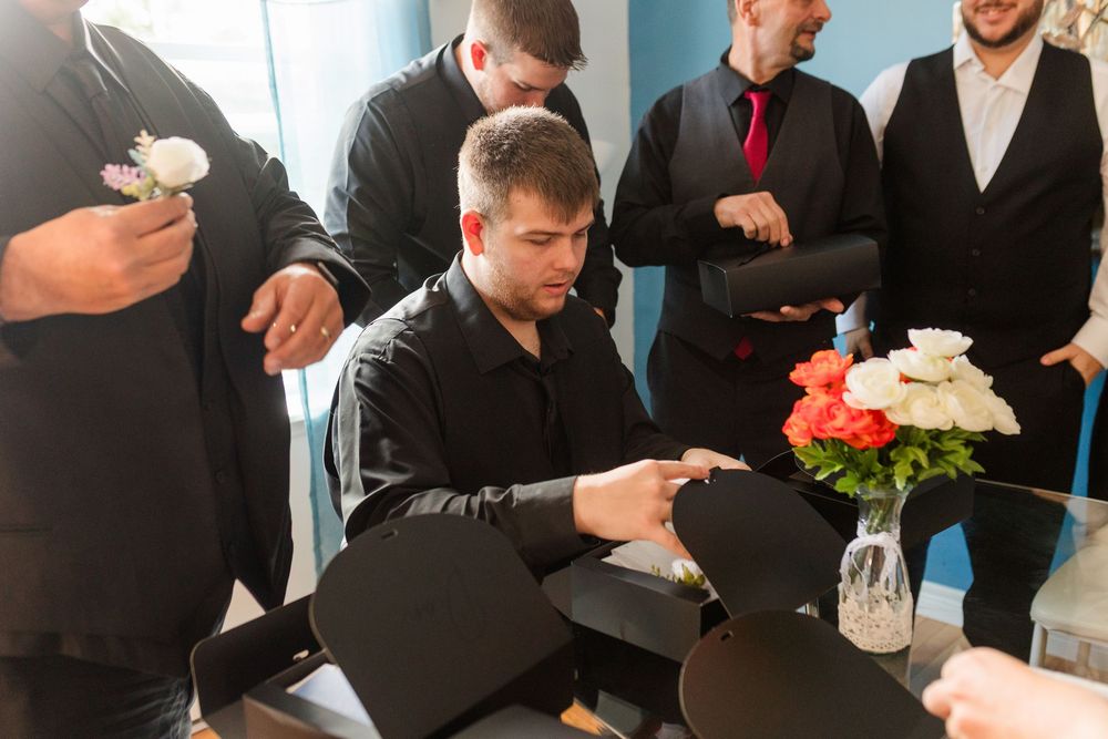 A group in formal black attire gathers around a table with flowers in a vase during what appears to be a business meeting.