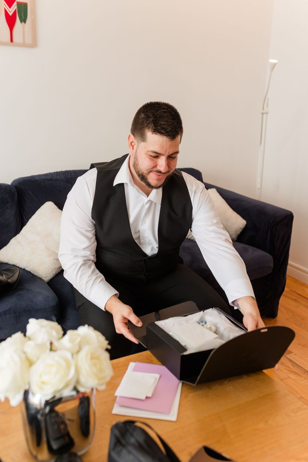 A person in business attire sits on a dark couch looking at documents in a box near white roses and notebooks.