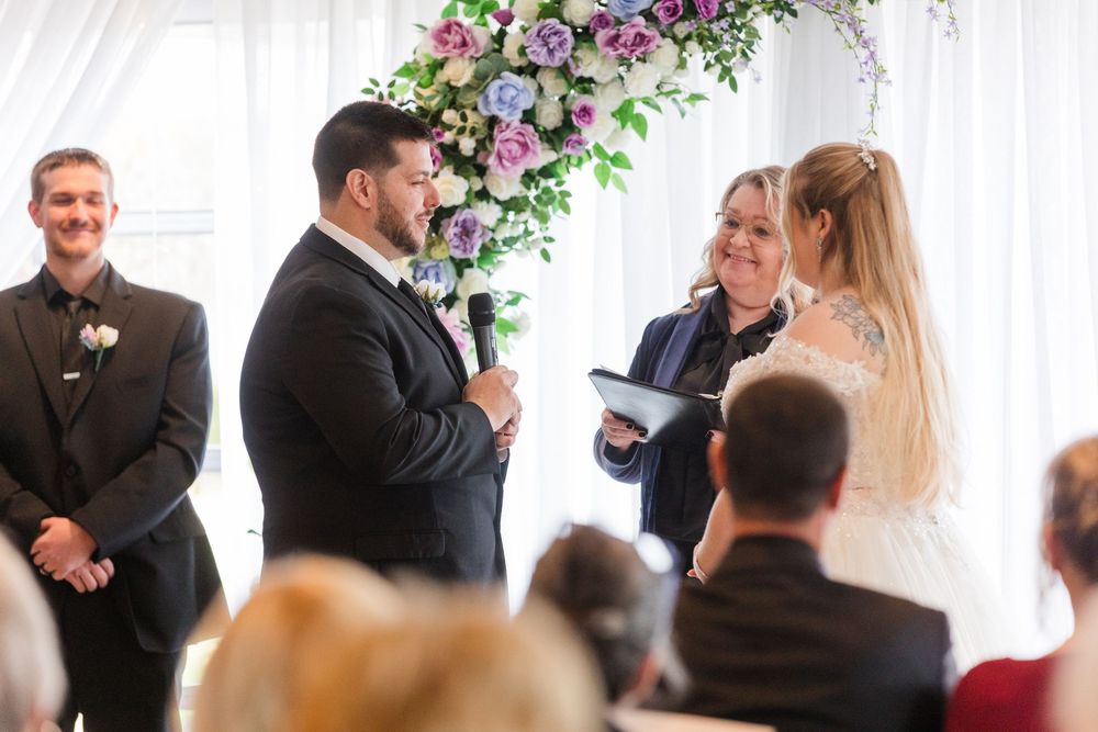 Wedding ceremony guests watch as bride and groom exchange vows under floral arch during indoor celebration.