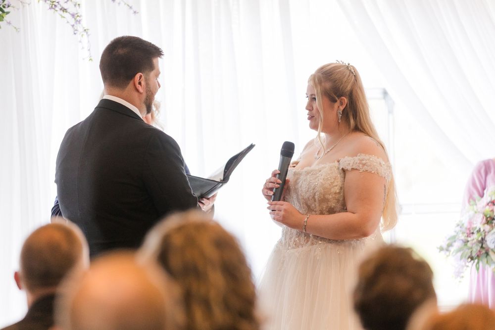 Wedding guests listen attentively during an indoor reception ceremony with pink floral decorations.