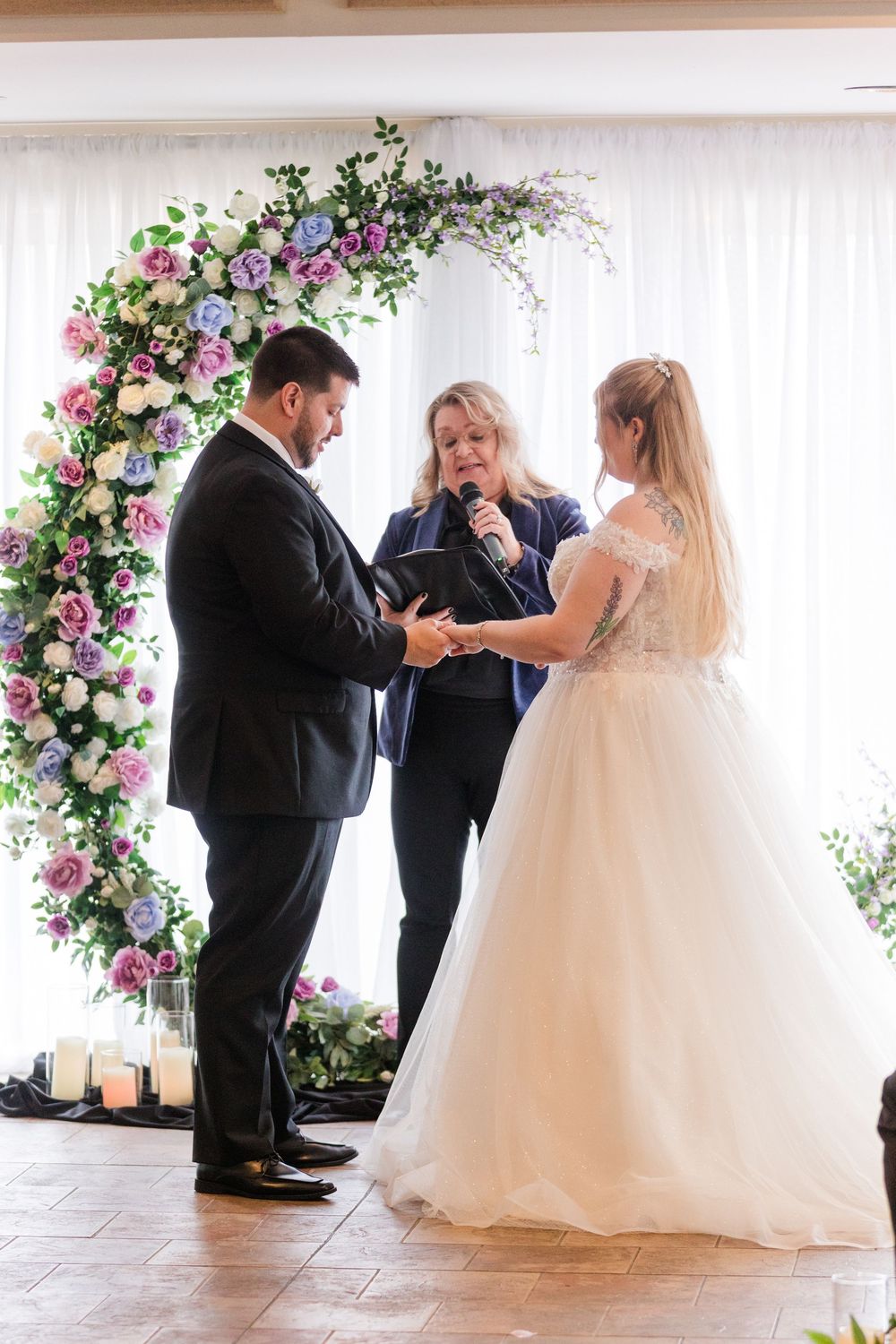 Emotional wedding ceremony moment under floral arch with bride in white gown and two others sharing heartfelt exchange.