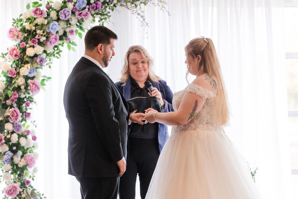 A wedding ceremony takes place under a floral arch with pink and white roses.