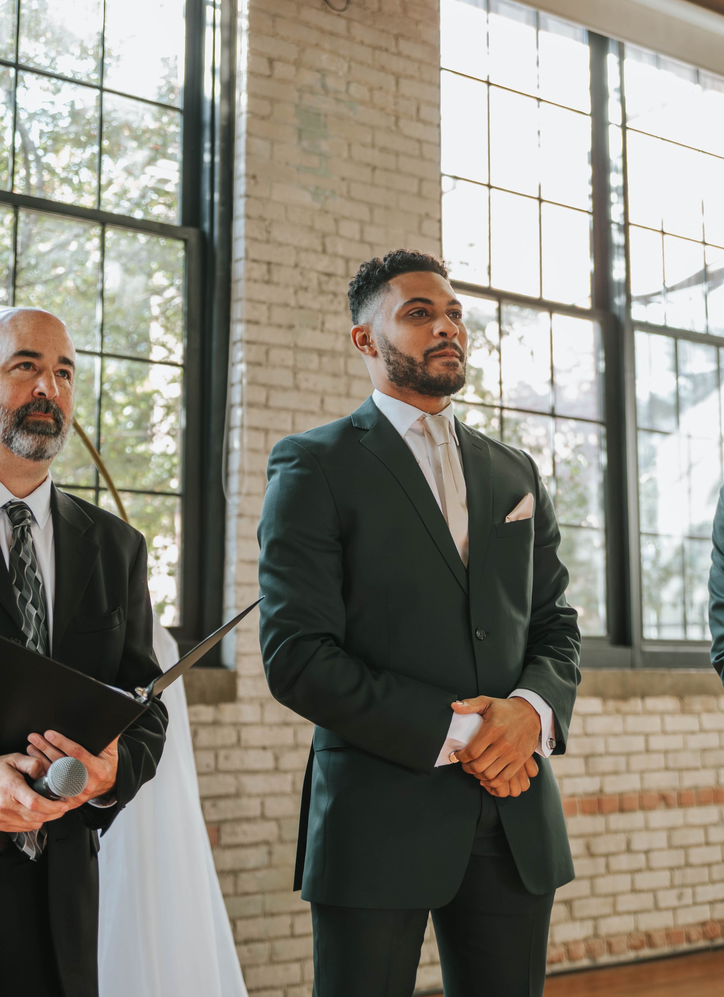 Two men in formal dark suits stand in an elegant venue with white brick walls and large windows.